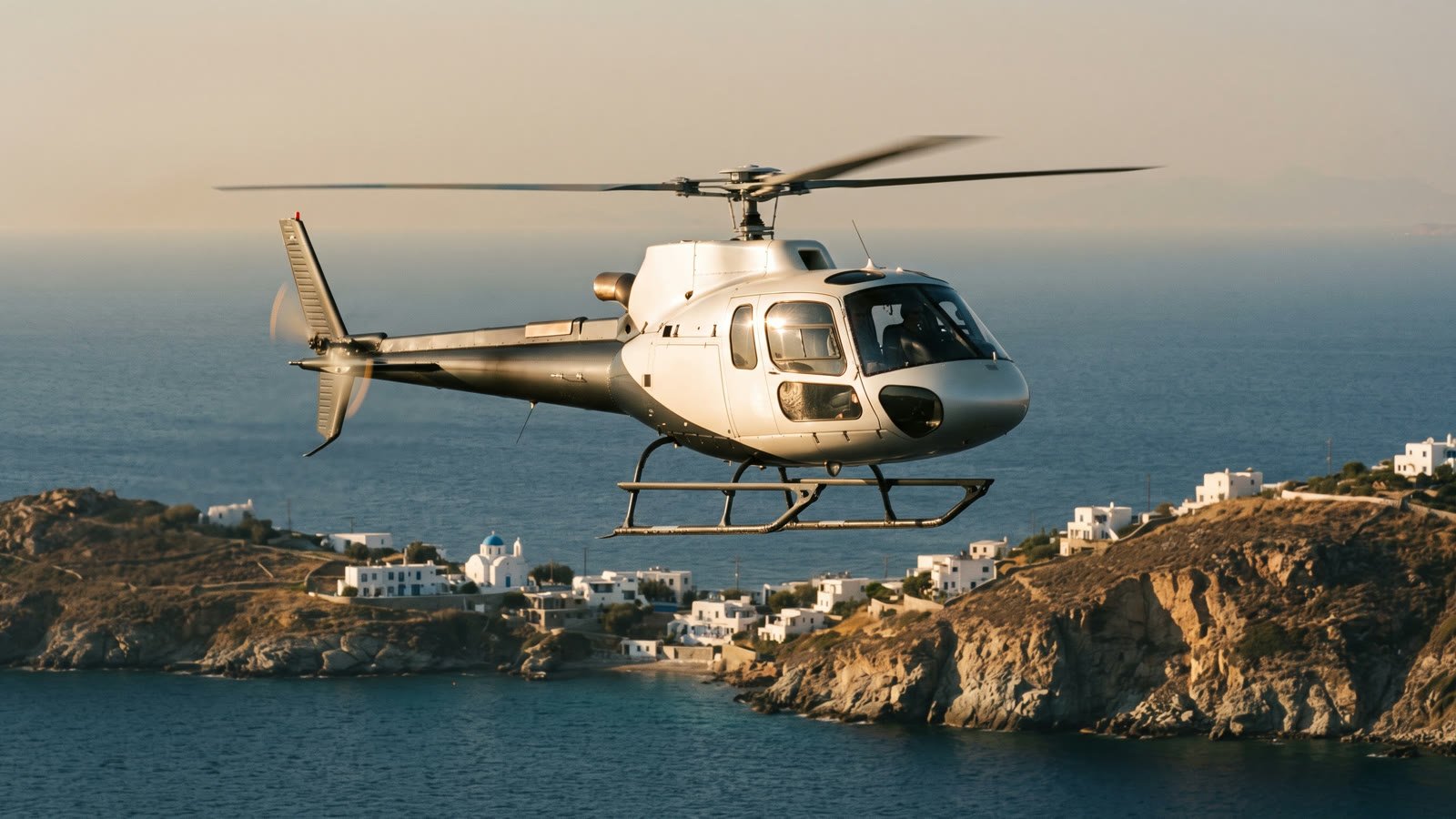 Private charter helicopter flying low over the deep blue Aegean at golden hour, a whitewashed Cycladic village with a blue-domed church on the coastline below