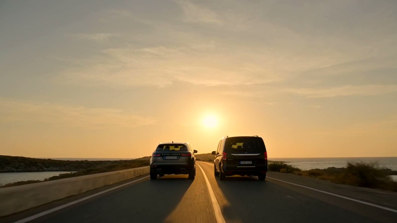 Range Rover Velar and Mercedes V-Class driving together into a golden Paros sunset on a coastal road